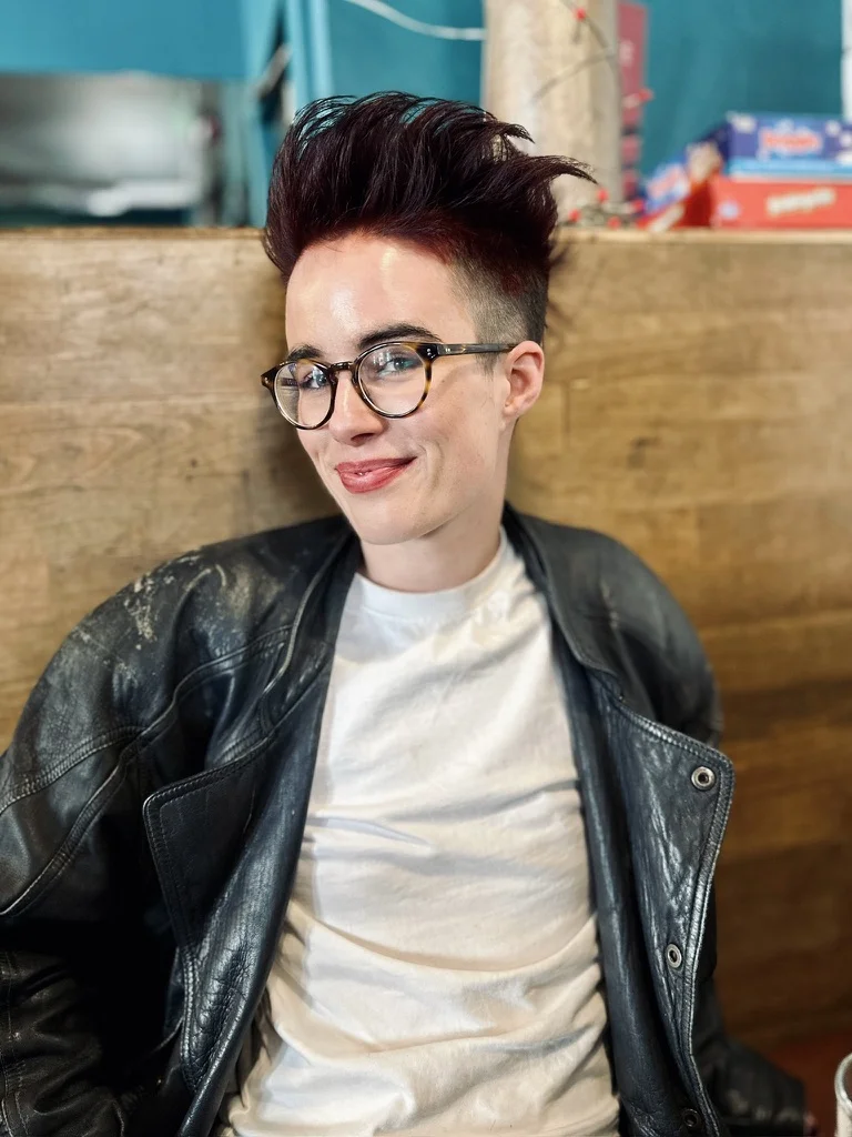 Lou, in tortoiseshell glasses and a black leather jacket, sitting in a café booth